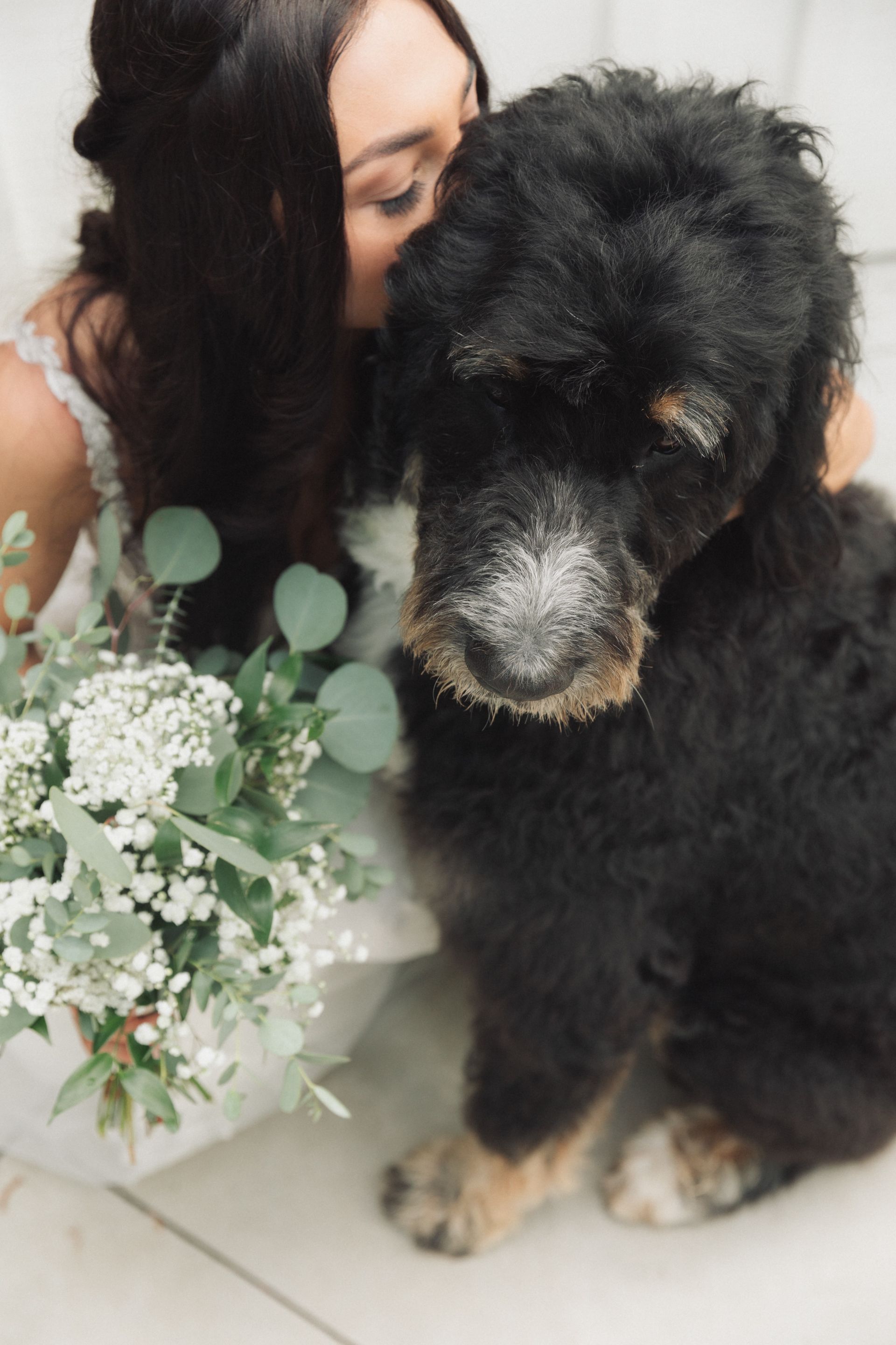 A bride is kissing a small black dog while holding a bouquet of flowers.
