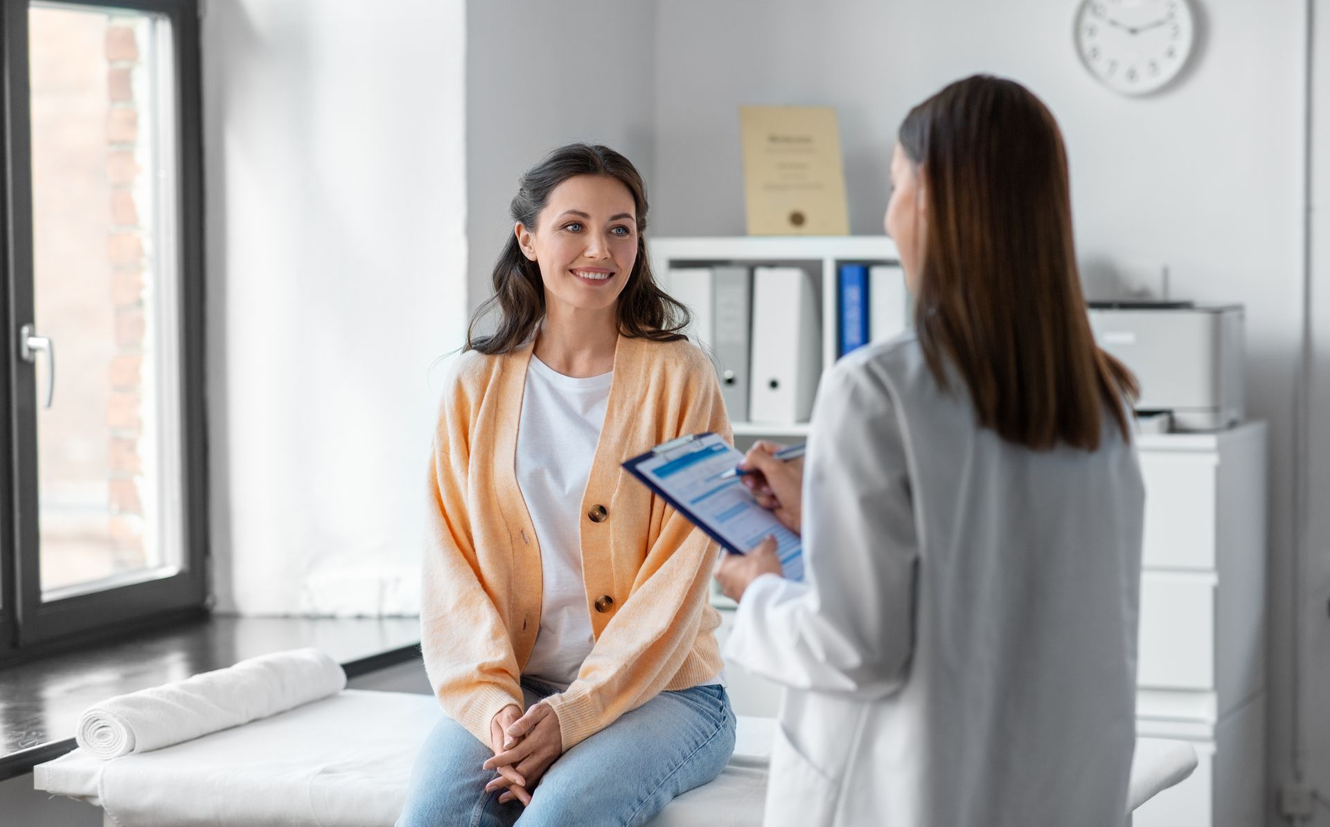 Person seated on an exam table consulting with a healthcare professional holding a clipboard.