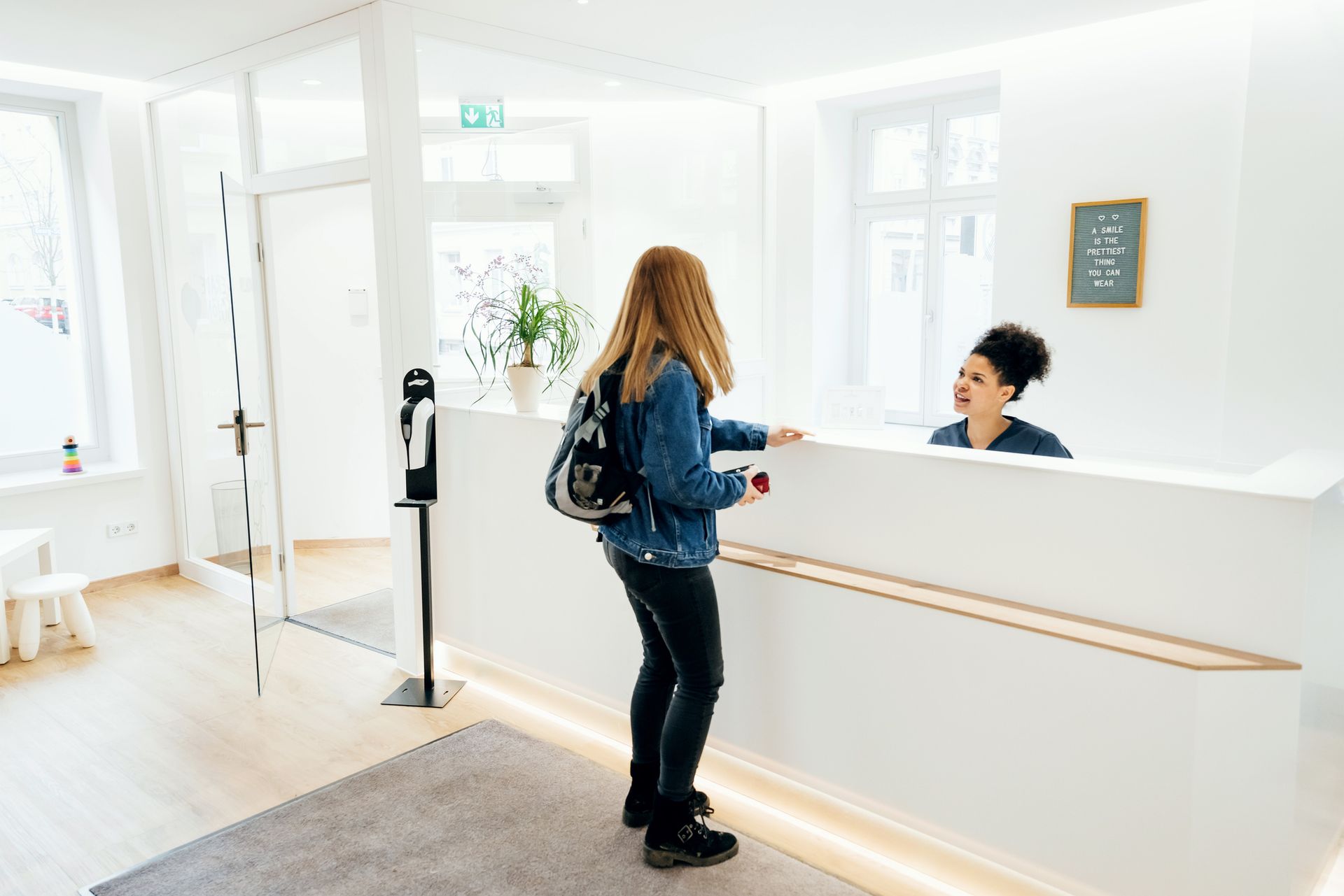 Woman at reception desk, speaking to receptionist in white waiting area.