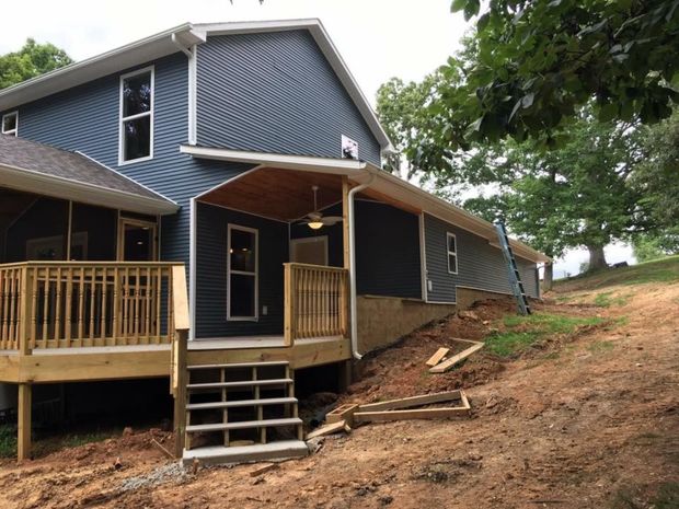 Blue house with wooden porch on a sloped dirt hillside under trees