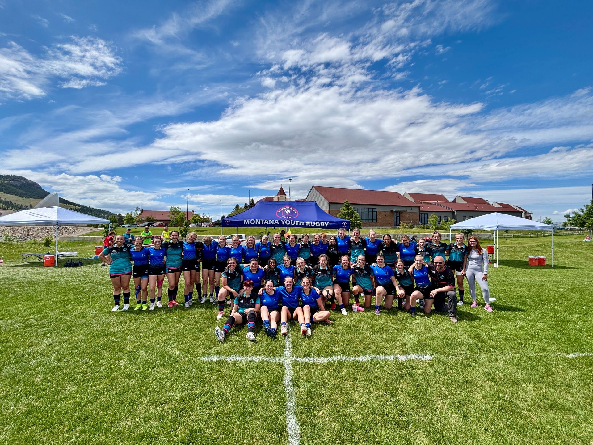 Group of young people in blue and teal uniforms on a grassy field with buildings and blue sky.
