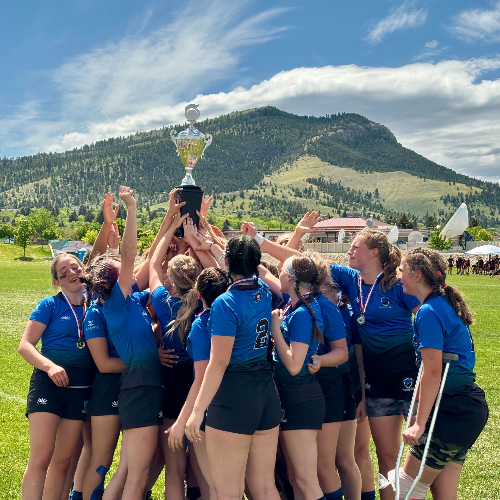 Women's rugby team celebrating a victory, holding trophy. Mountain backdrop, sunny day.