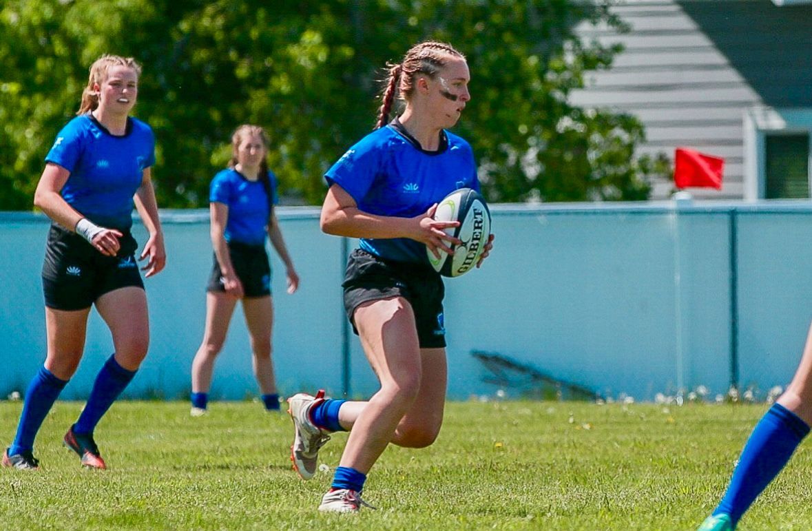 Female rugby players in blue jerseys on a sunny field, one running with the ball.