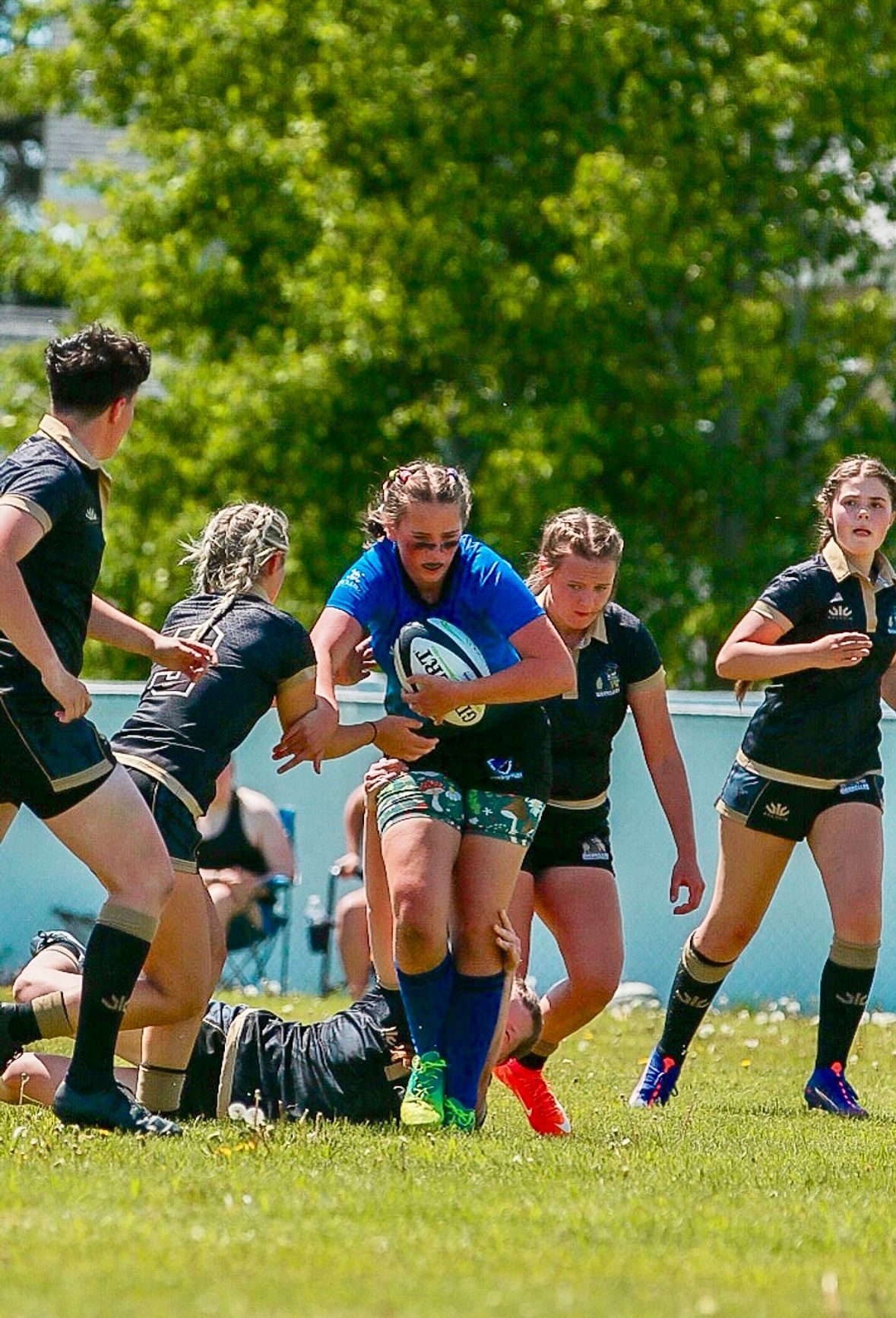 Woman in blue shirt runs with rugby ball; players in black attempt to tackle.