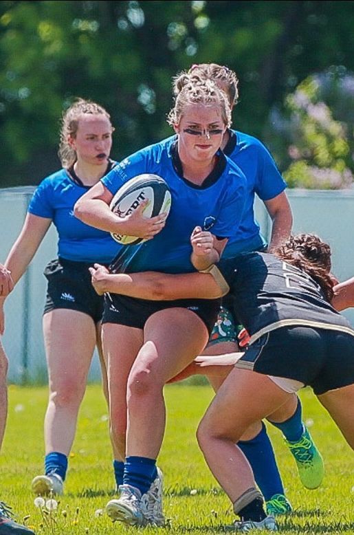 Female rugby player in blue jersey runs with ball, being tackled by opponent in black jersey on green field.