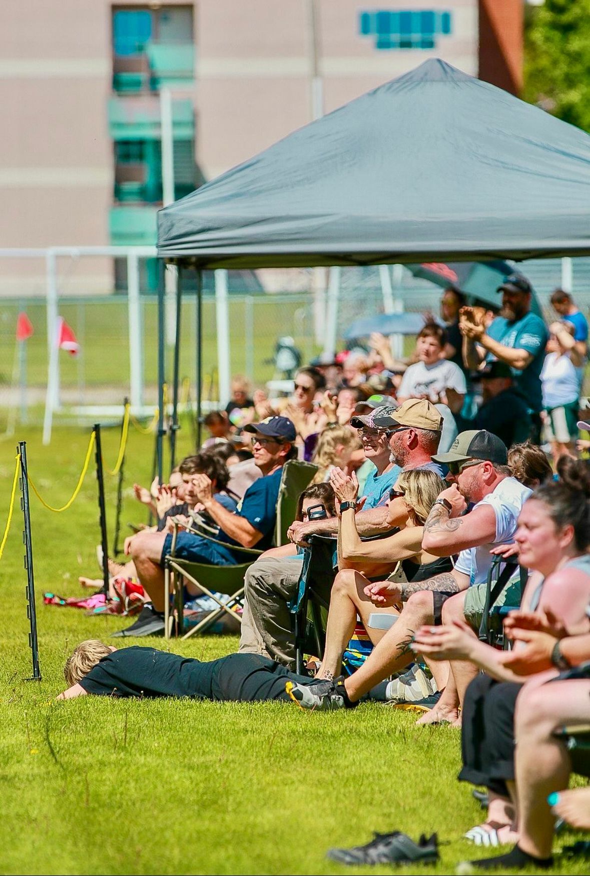 Spectators watching an outdoor event, sitting on the grass under a canopy. Sunny day, green field.