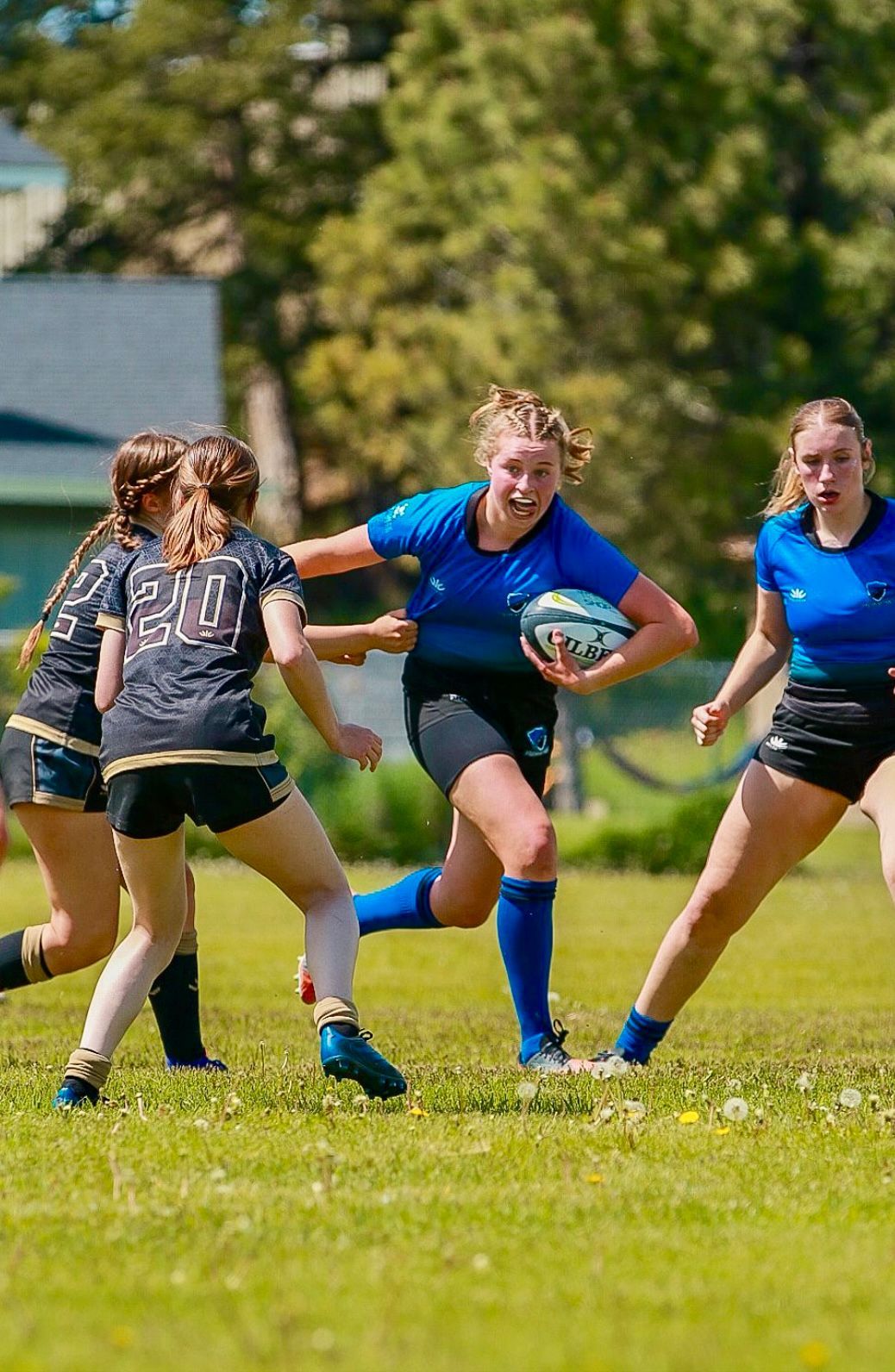 Female rugby player in blue jersey runs with ball, pursued by two opponents on grassy field.