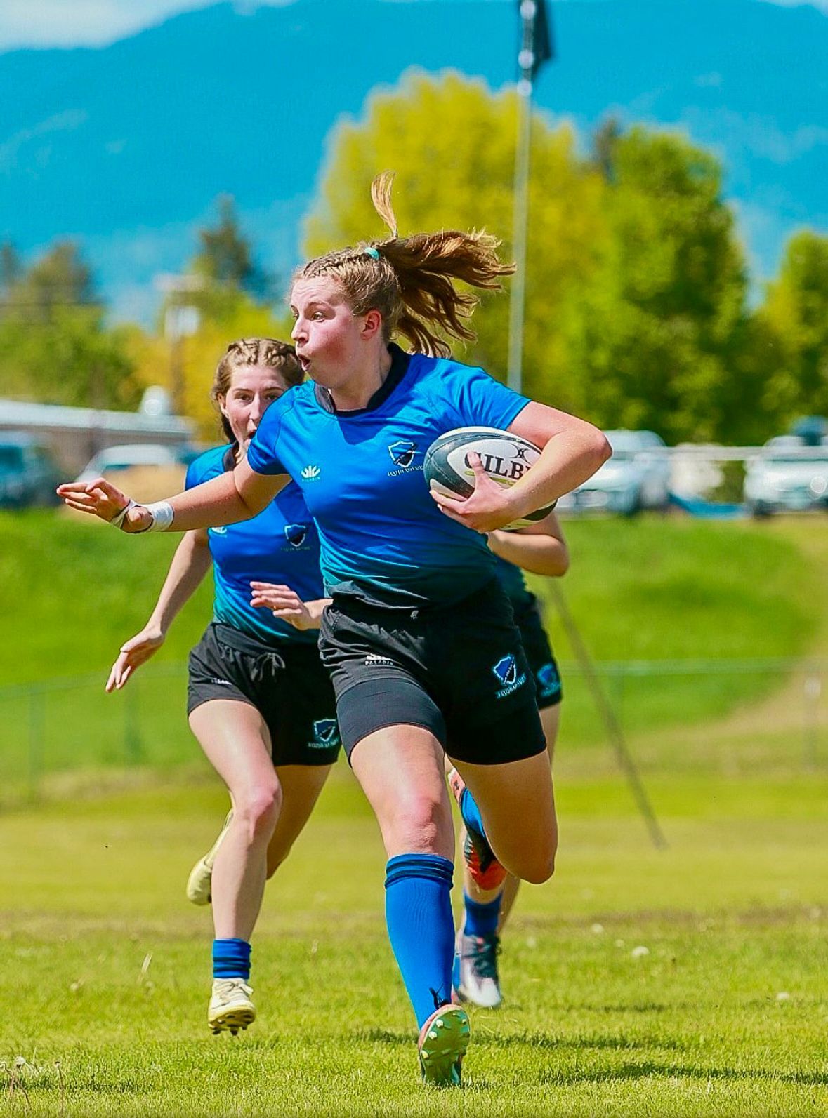 Woman in blue rugby uniform runs with ball on grass field, mountains in background.