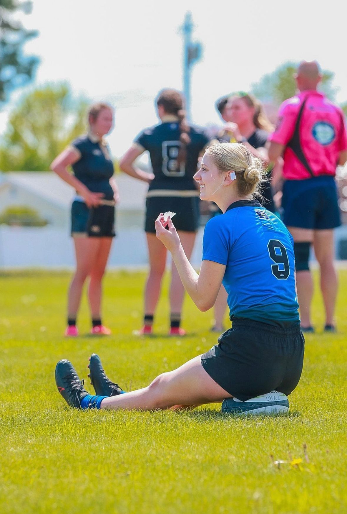 Woman in blue jersey, sitting on field, talking. Teammates and referee in the background.