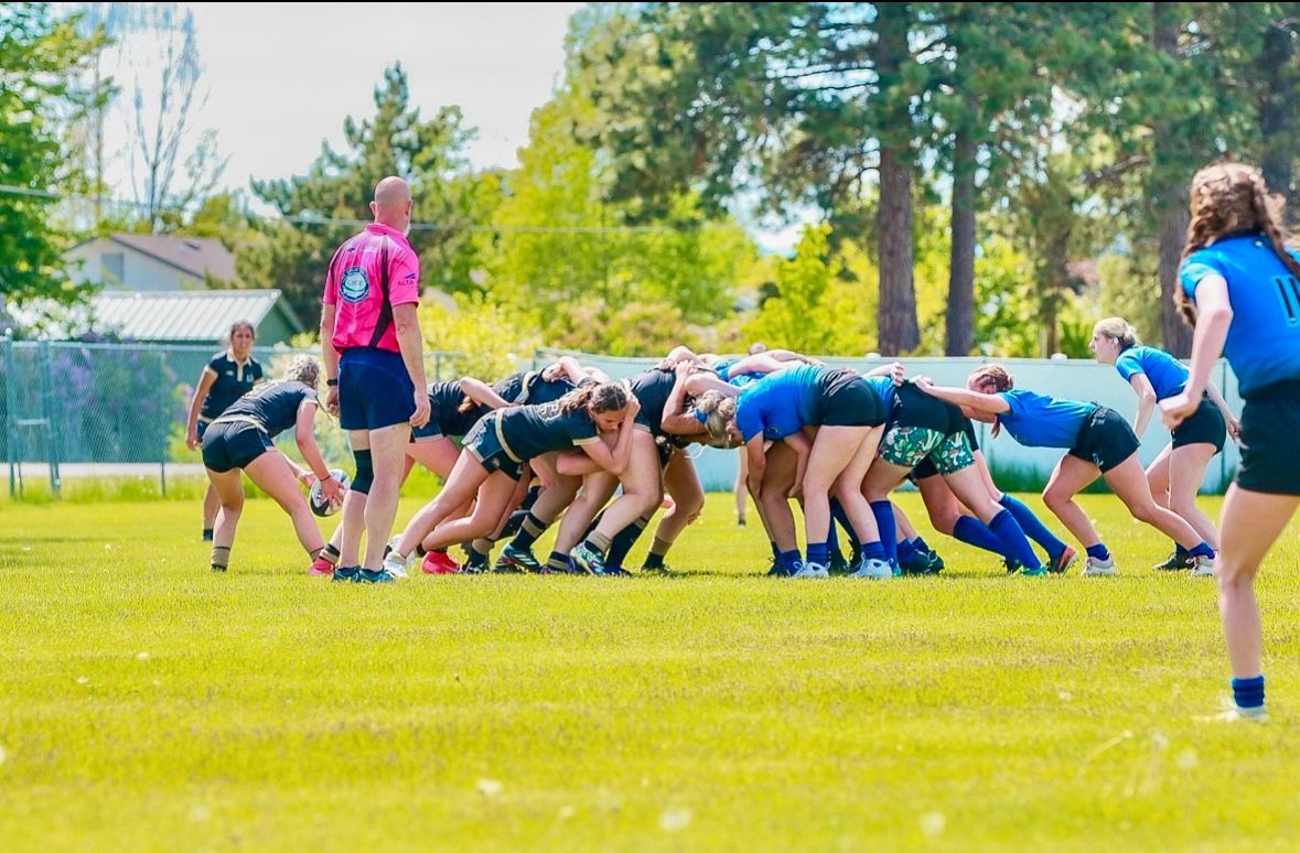 Women's rugby players in scrum on grassy field, referee watches. Teams in blue and black jerseys.