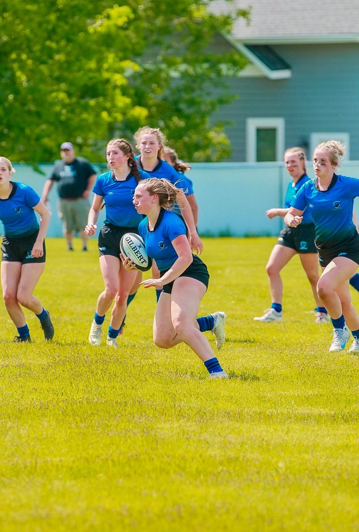 Women's rugby game: Player in blue jersey runs with ball, followed by teammates on a sunny field.