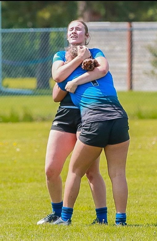 Two women athletes in blue jerseys hug on a grassy field.