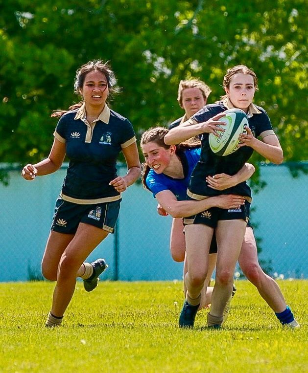 Women's rugby players in action: black uniforms, tackling on a green field, sunny day.