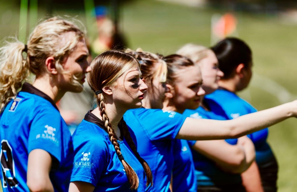 Women in blue sports jerseys stand on a field, one pointing, likely in a huddle or watching a game.