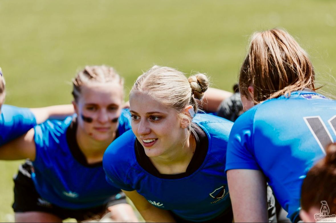 Female rugby players in blue jerseys huddle on a sunny field, smiling and focused.