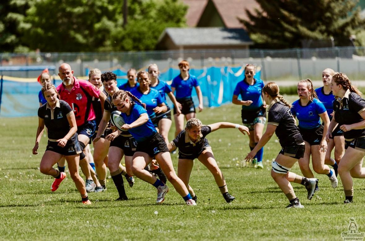 Women's rugby match: Players in blue and black uniforms run on a green field under a sunny sky.
