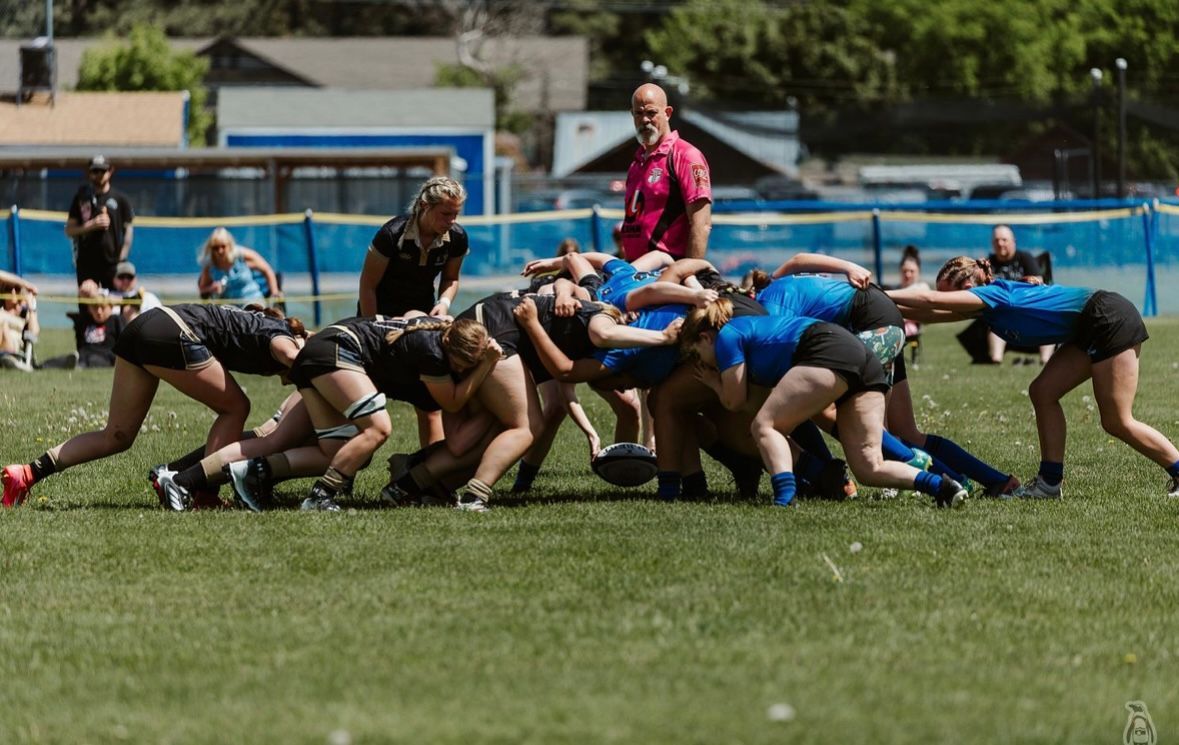 Women's rugby players in black and blue jerseys engage in a scrum on a grassy field.
