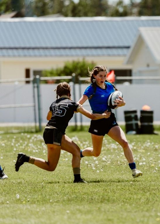 A rugby player in blue runs with the ball, pursued by a player in black, on a grassy field.
