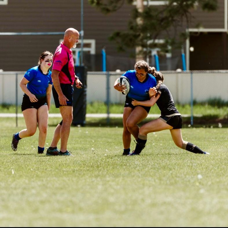 Female rugby player in blue jersey tackled on a green field. Referee watches, another player runs.