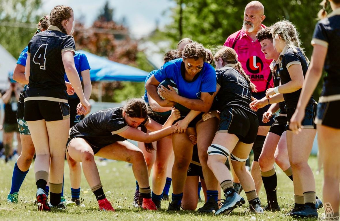 Rugby players in action, tackling on a grassy field; referee watches.