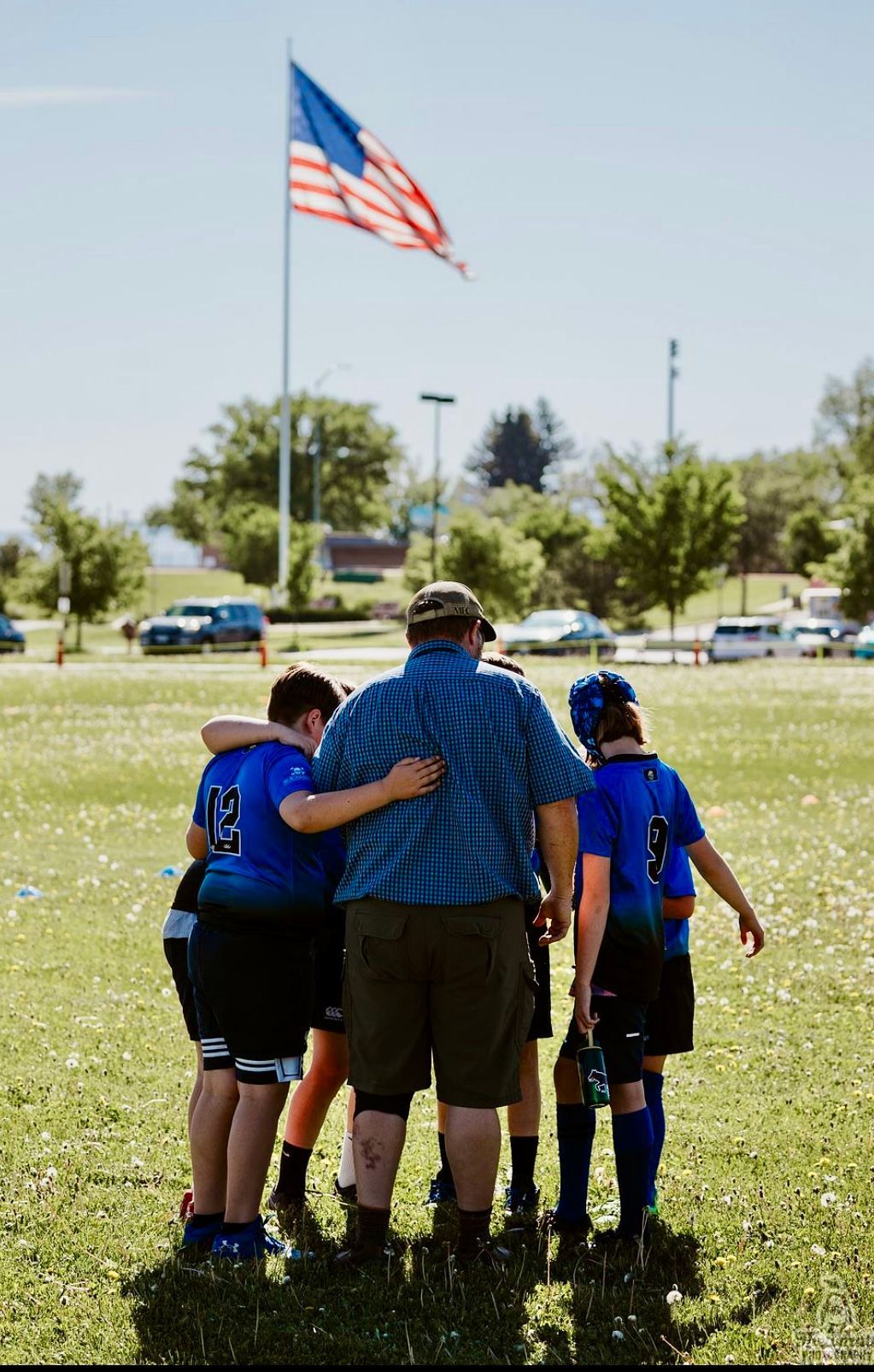 A coach and team huddle on a grassy field under the American flag. They wear blue uniforms.