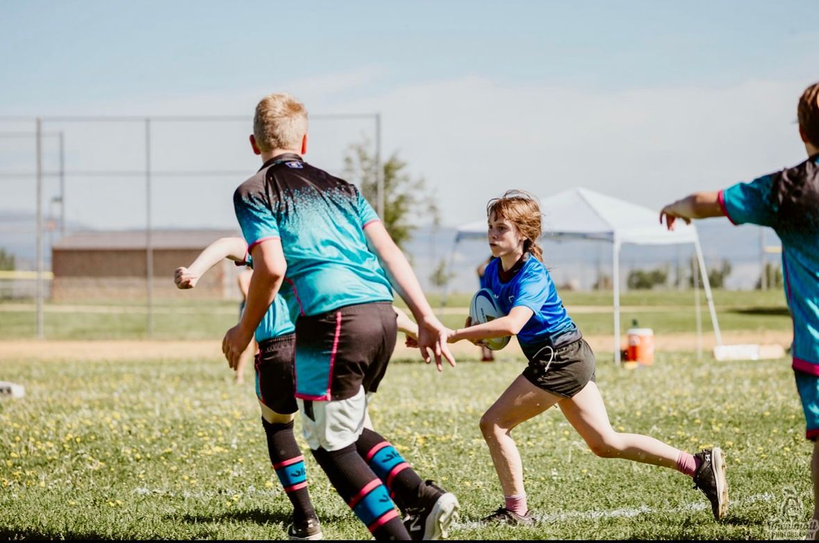 Children playing rugby on a grassy field, one running with the ball, wearing blue jerseys.