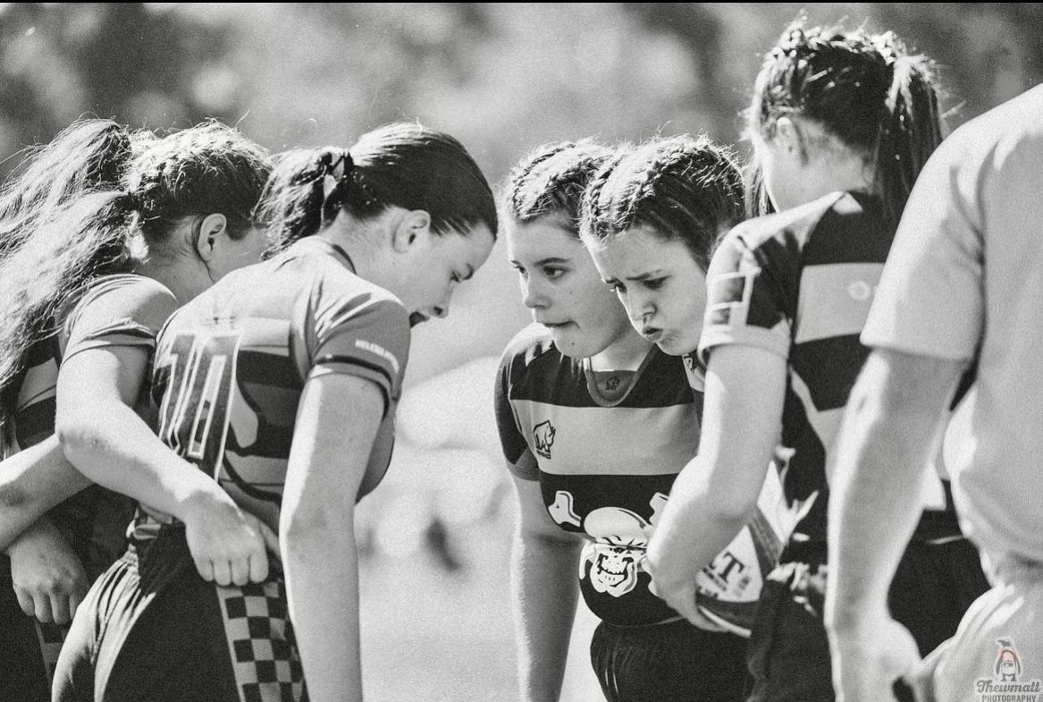 Female rugby players in a huddle, focused and determined, on a sunny field. Black and white.