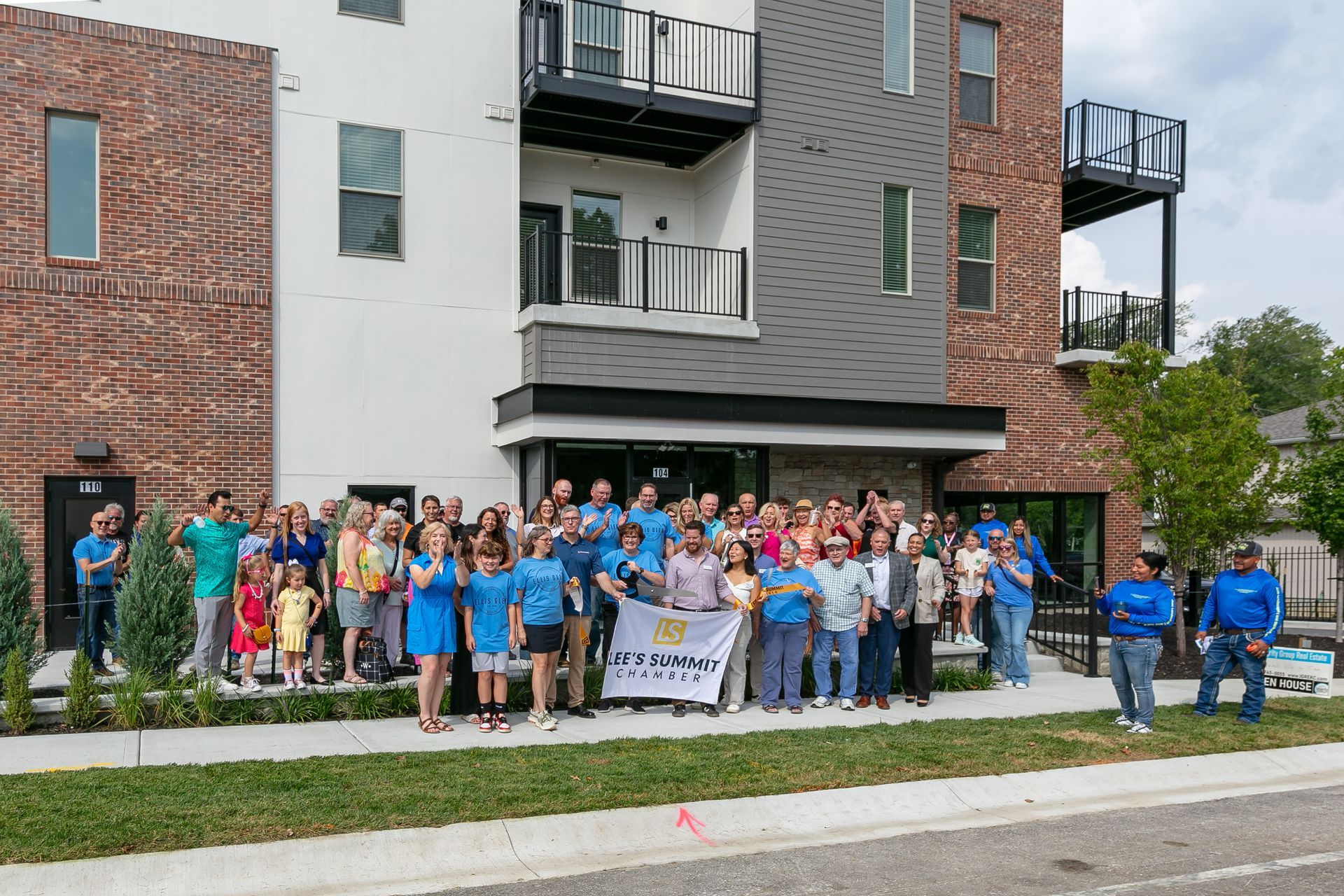 Group of people in front of a building holding a banner. Many are wearing blue. Sunny day.