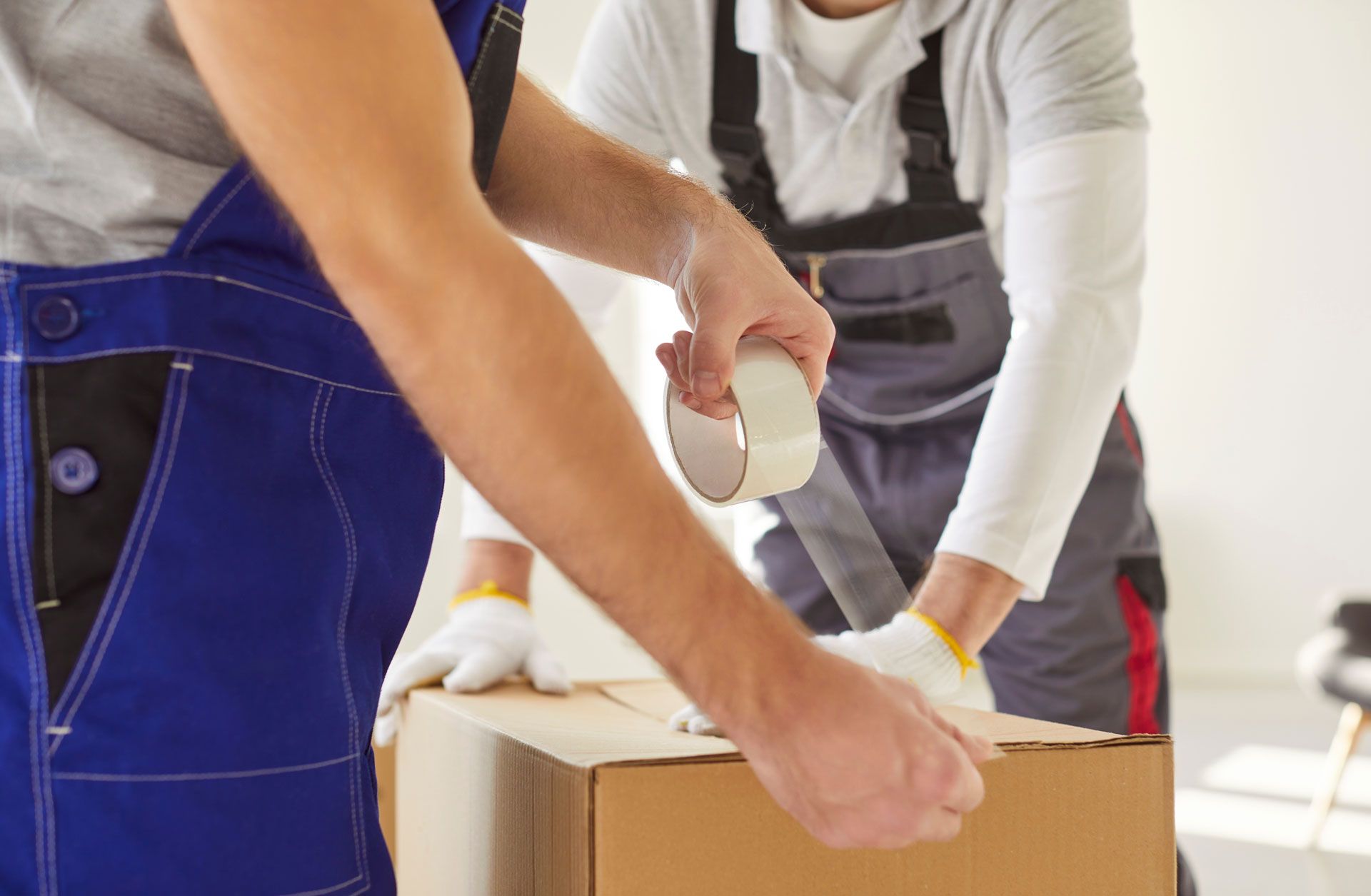 Two people packing a cardboard box with tape, wearing work overalls and gloves.