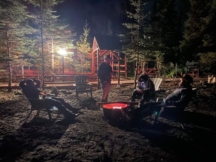 People sitting around a glowing campfire at night near a lit playground in the woods