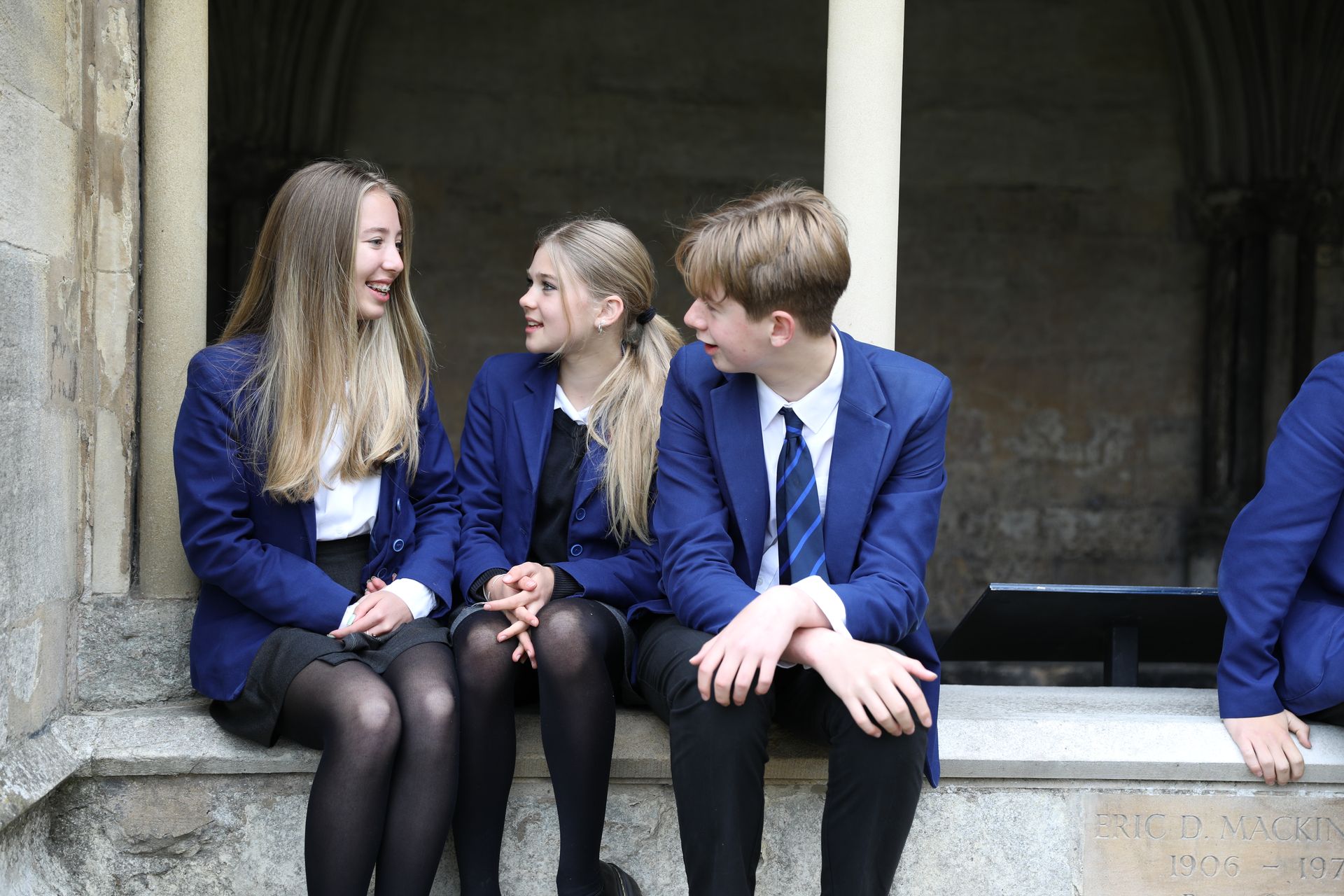 A group of young people in school uniforms are sitting on a ledge talking to each other.