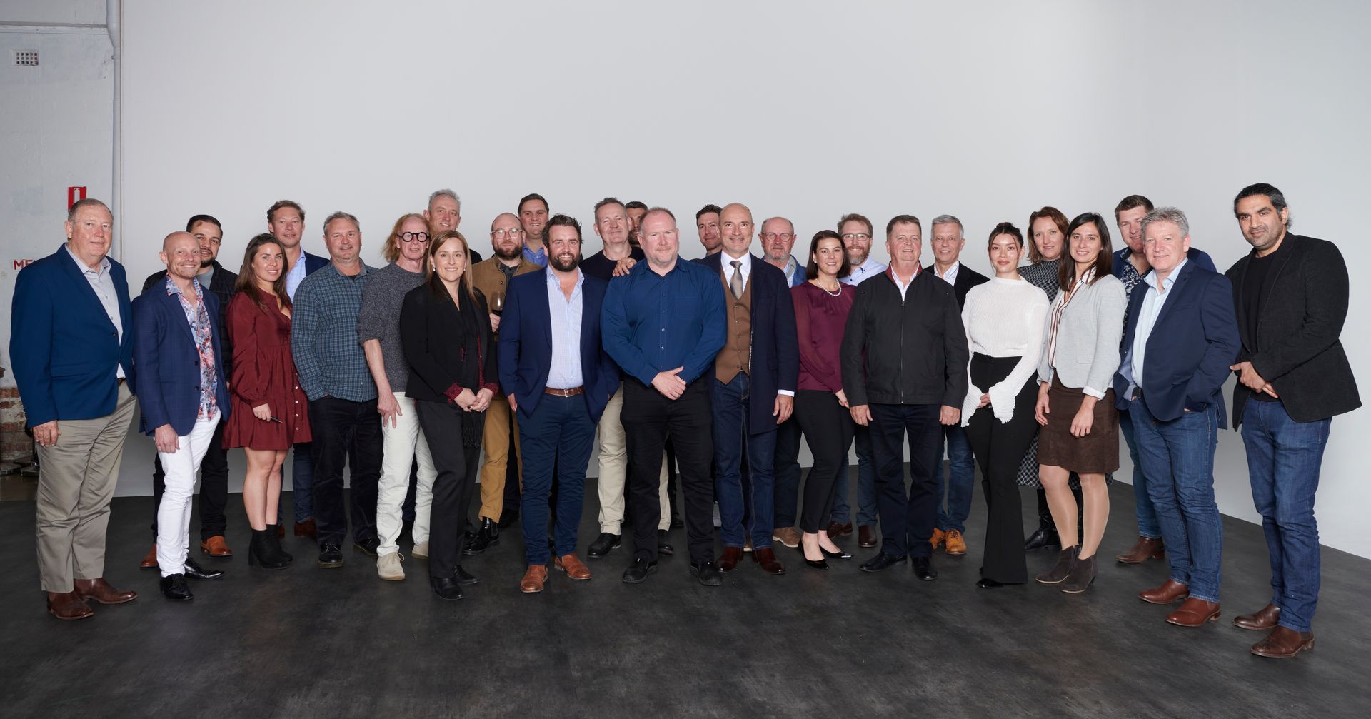 Group of people posing in front of a white wall. Men and women dressed in business casual attire.