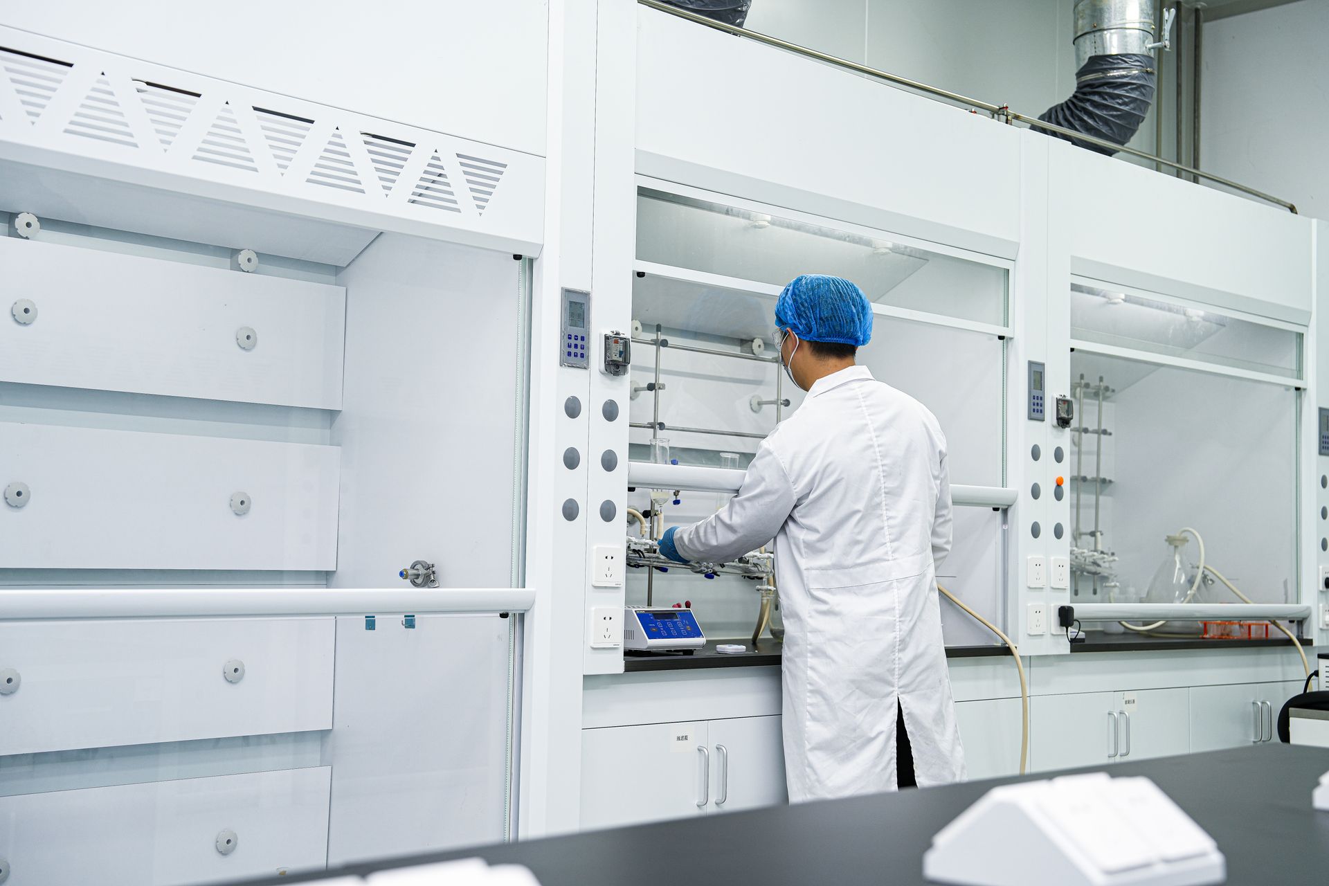 Scientist in lab coat and hairnet working with equipment inside a fume hood in a laboratory.