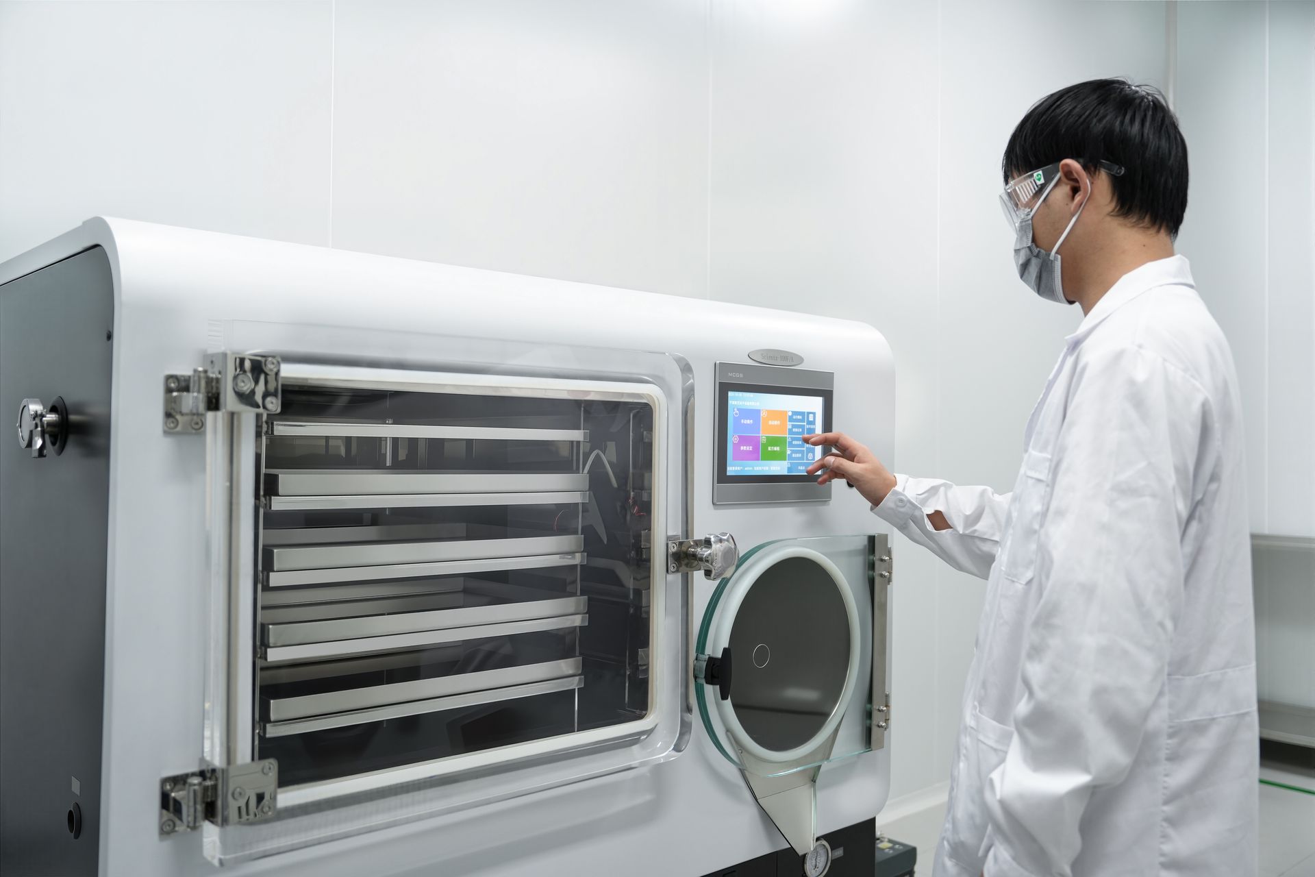 Man in lab coat operates freeze dryer in a white lab, touching the machine's touchscreen control panel.