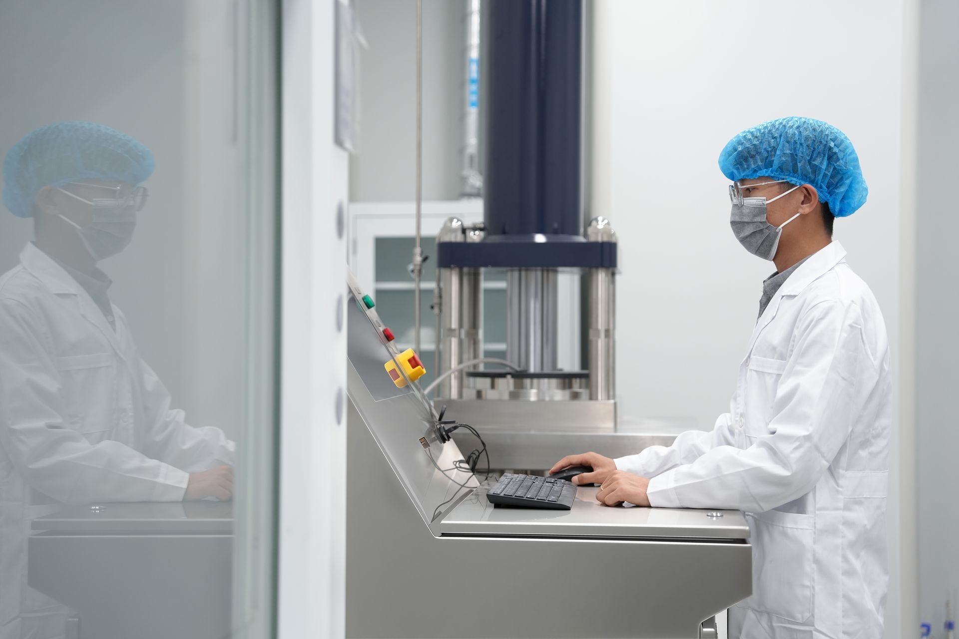 Person in lab coat and hairnet operating machinery in a laboratory; reflection visible.