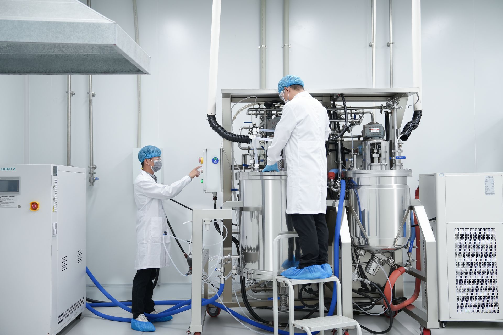 Two people in lab coats work with machinery in a cleanroom. One adjusts controls, the other stands on a step stool.