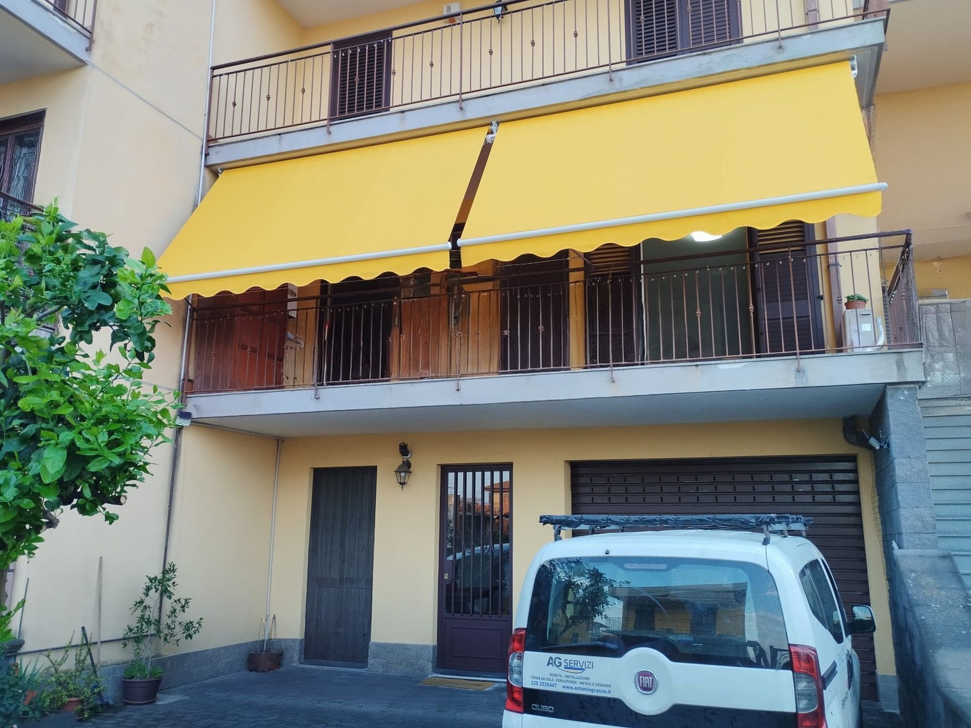 Tenda gialla sulla facciata di un condominio con balconi e un furgone bianco parcheggiato sotto.