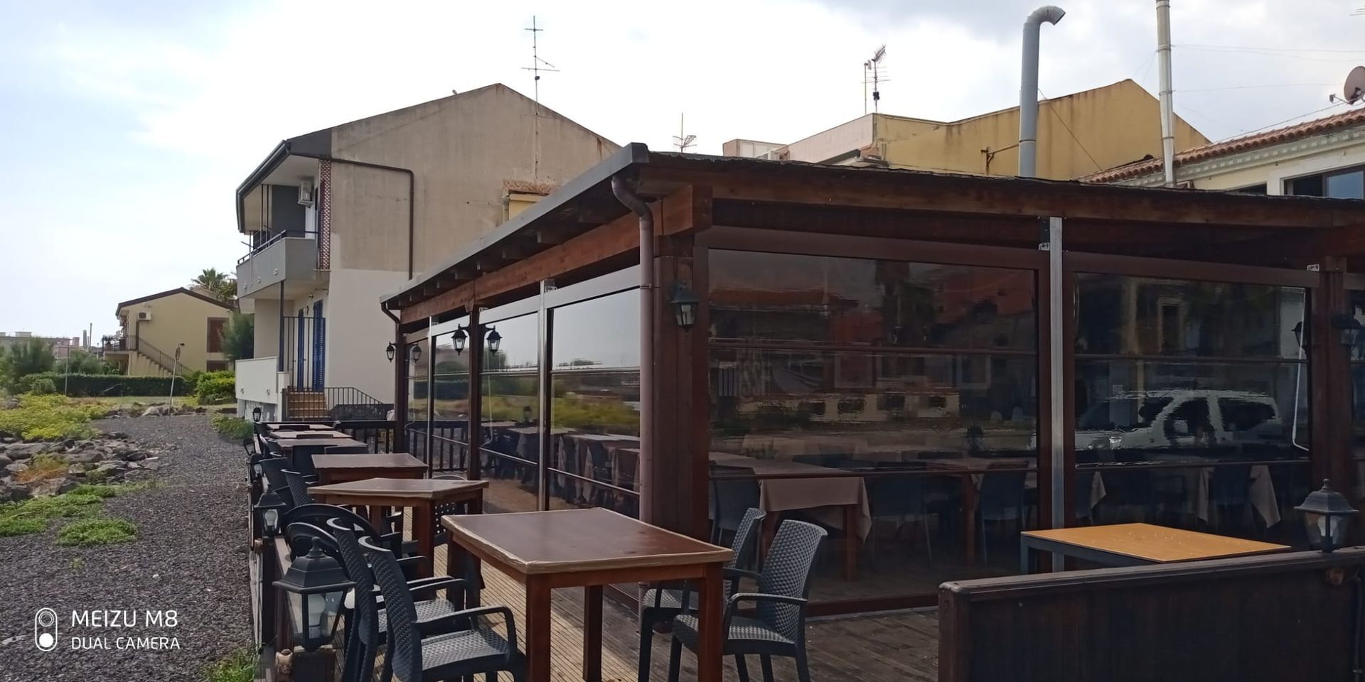 Terrazza esterna del caffè con tavoli e sedie in legno, adiacente agli edifici di una strada soleggiata.