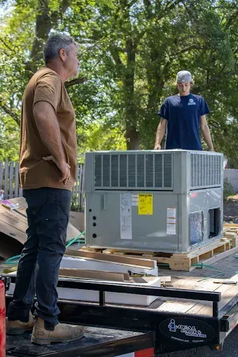 Two workers stand on a flatbed trailer next to a new, large gray HVAC unit on a wooden pallet outdoors.