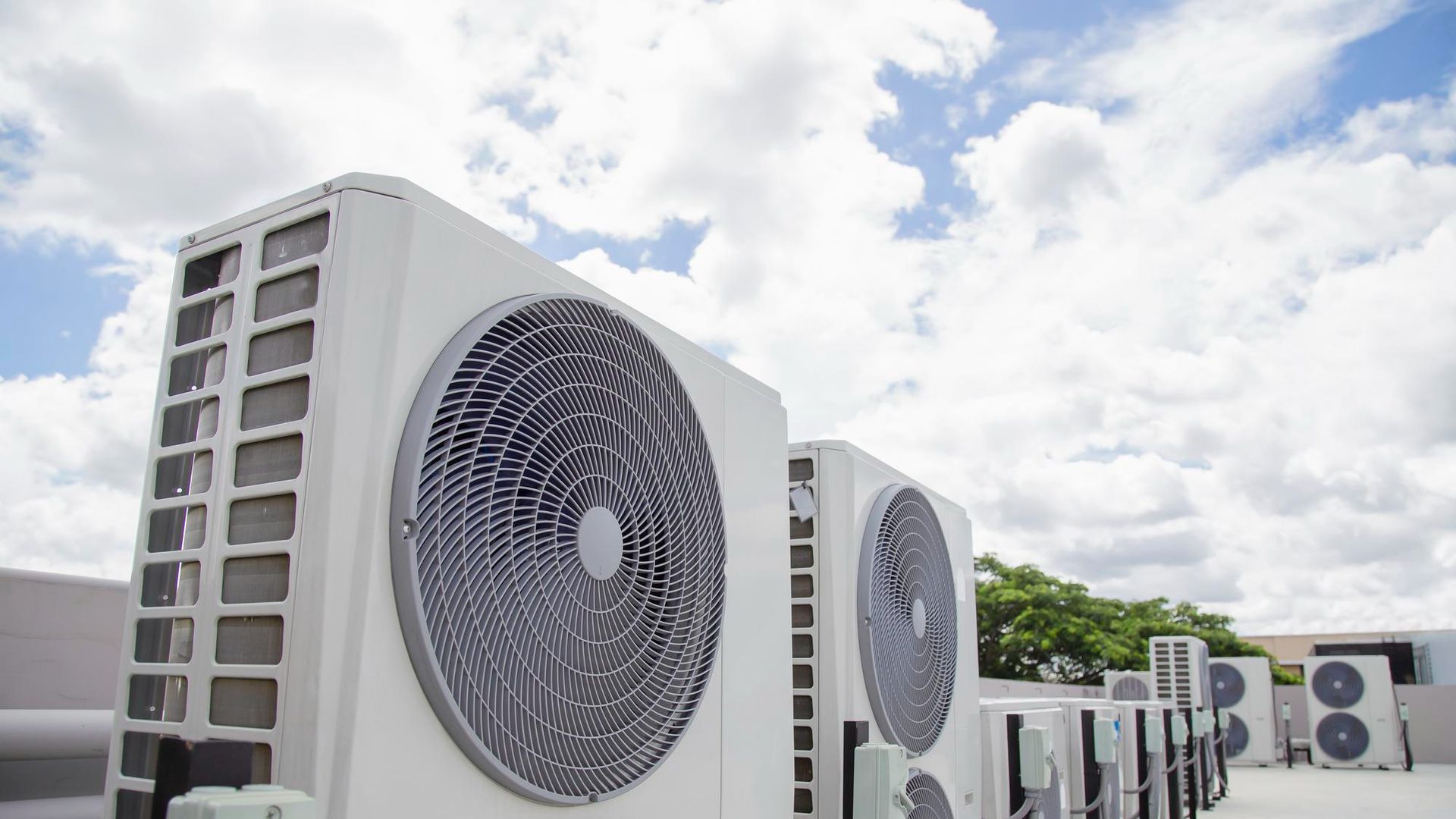 A row of white industrial HVAC air conditioning units on a rooftop under a bright, cloudy sky.