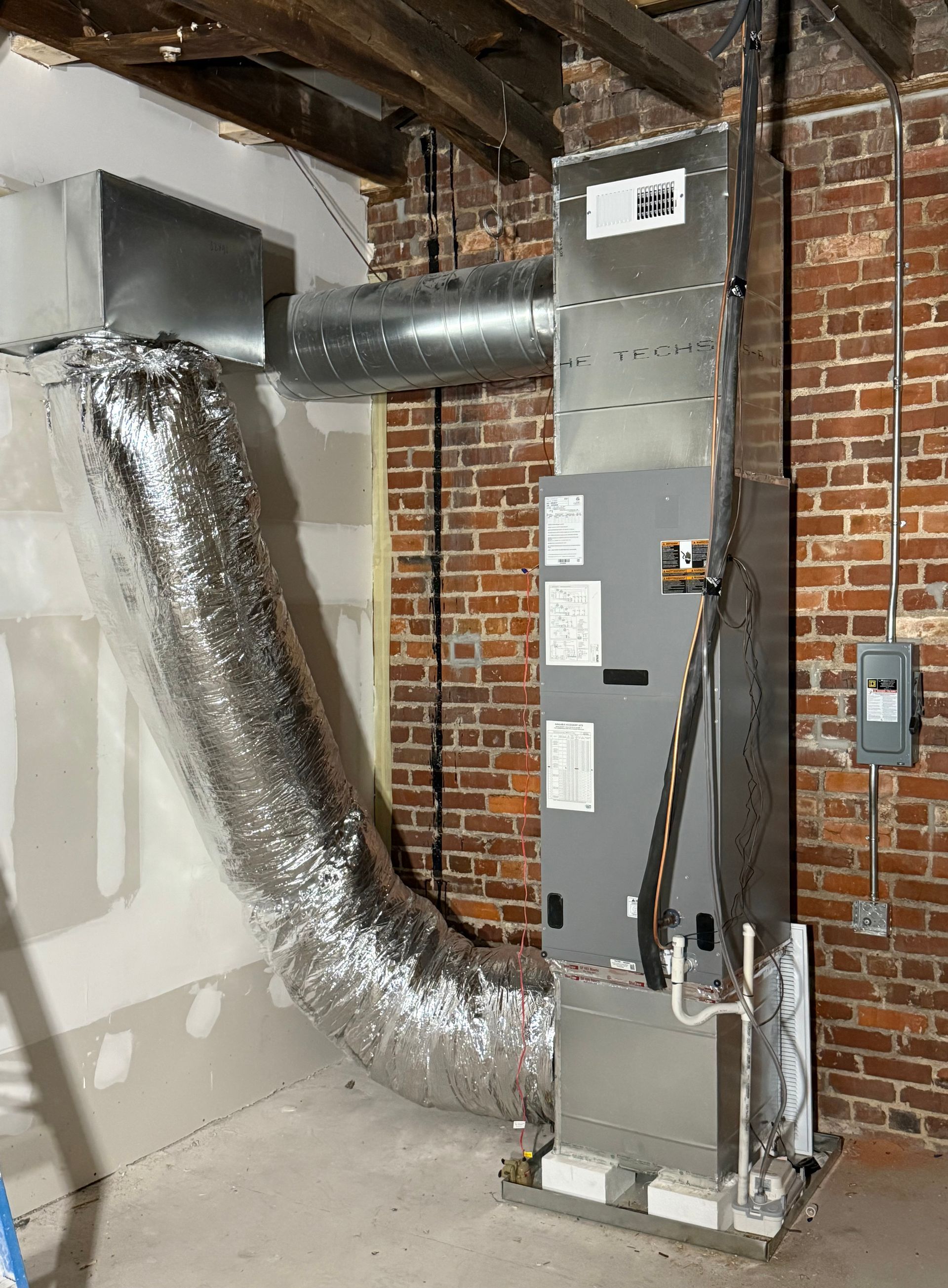 An HVAC air handler unit standing against a brick wall, connected to ductwork in an unfinished basement.