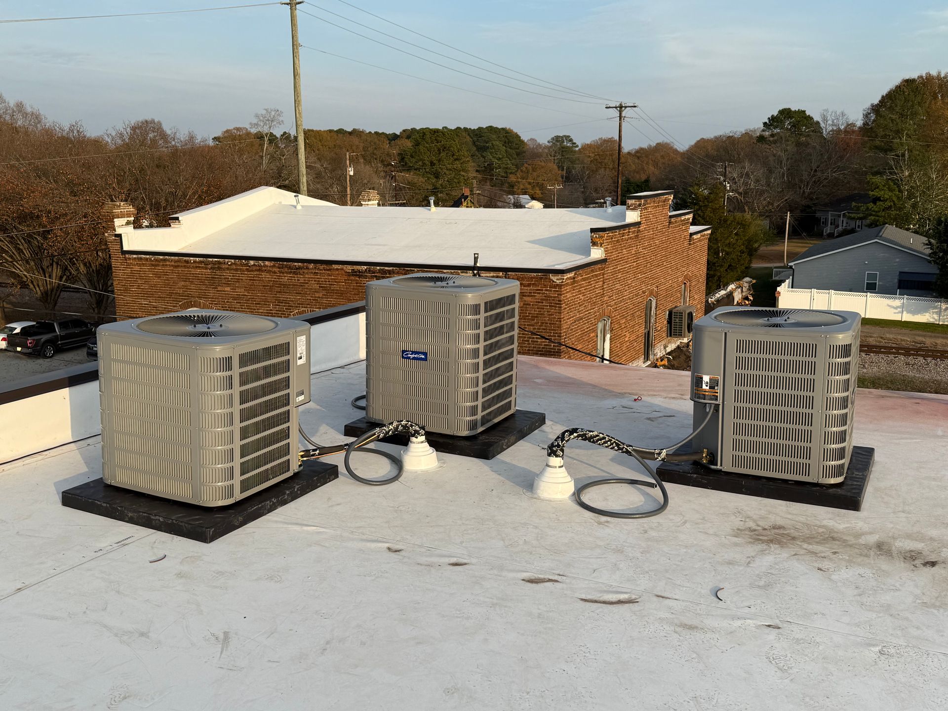 Three air conditioning units on a flat white commercial roof with a brick building in the background.