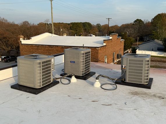 Three air conditioning units on a flat white commercial roof with a brick building in the background.