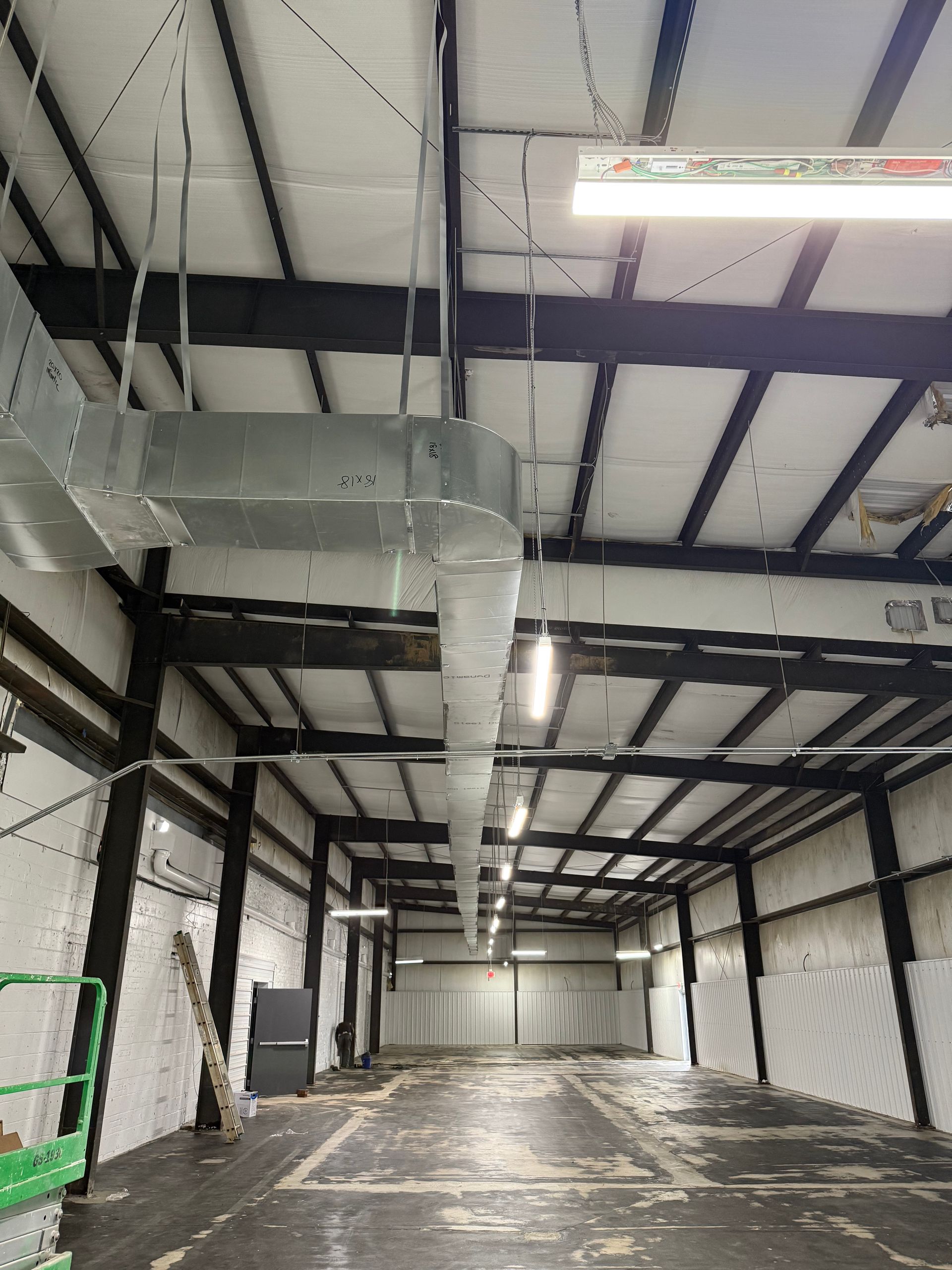 A long, empty industrial warehouse with dark metal rafters, white walls, and a large suspended metal ventilation duct.