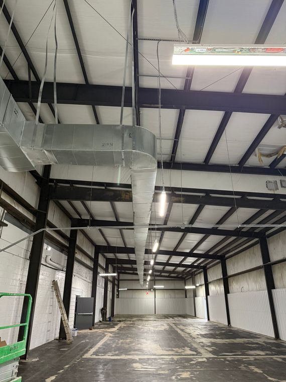 A long, empty industrial warehouse with dark metal rafters, white walls, and a large suspended metal ventilation duct.