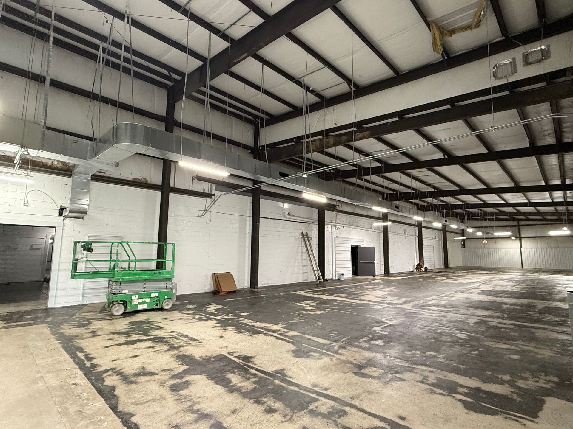 An empty industrial warehouse with a white wall, exposed ceiling beams, ductwork, and a green scissor lift on a dark floor.