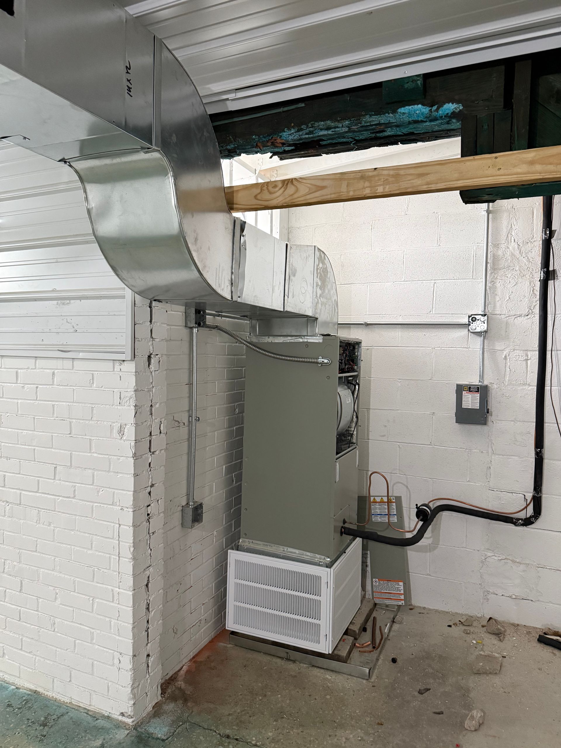 An HVAC air handler unit with attached ductwork stands in a basement against a white-painted brick wall.