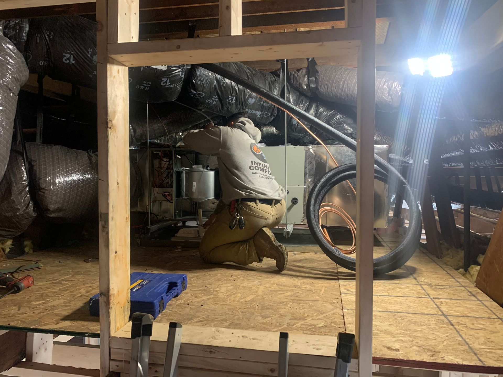 A professional in a gray shirt and tan pants kneels in an attic, working on HVAC equipment surrounded by insulation.