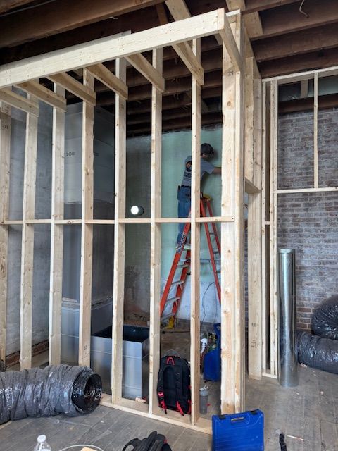 A person on a ladder works inside a wooden wall frame in a room with brick walls and exposed HVAC ductwork.