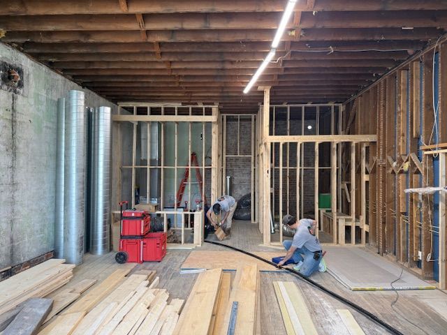 Two workers construct wooden interior wall frames inside a building under renovation with exposed ceiling beams.